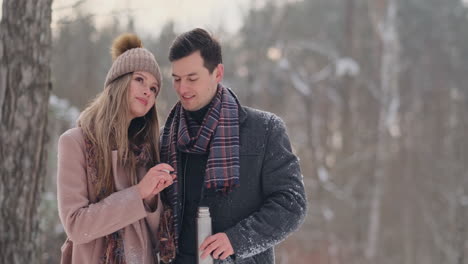 Happy-couple-holding-hot-tea-cups-over-winter-landscape.-Young-couple-in-love-on-a-winter-vacation,-standing-next-to-a-tree-and-drinking-hot-cup-of-tea